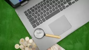 Na imagem, um notebok aberto com uma lupa próxima ao mouse e moedas com notas ao lado. Apostas esportivas online.